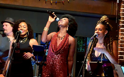 Three women in evening gowns singing into microphones. A man in a fedora is in the background.