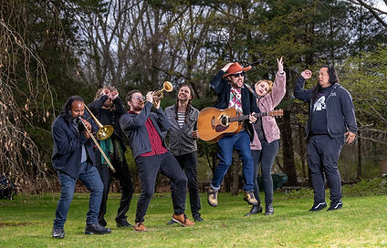 Group of casually-dressed peple with in a tree-lined field playing instruments.