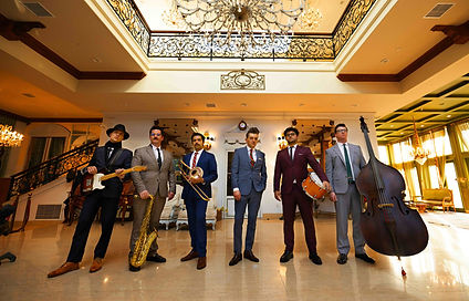 Group of men in tailored suits standing with instruments in an ornate ballroom.