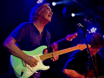 Close up of a man on stage singing with a green electric guitar. Another man can be seen in the backgrond.
