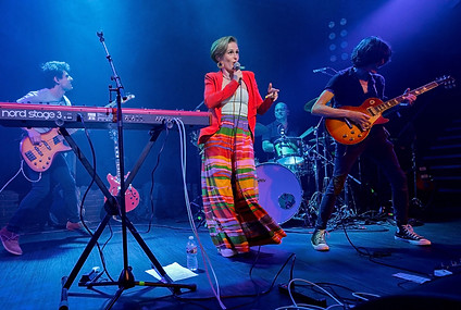 Woman in a bright, colorful outfit singing on stage with a full band behind her cast in blue light.
