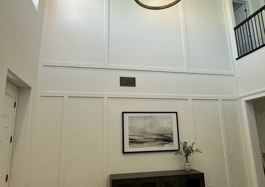 High-ceiling entryway with a double-height white board and batten feature wall. A dark charcoal console cabinet sits below framed black-and-white landscape art, completed by an open-ring chandelier overhead.