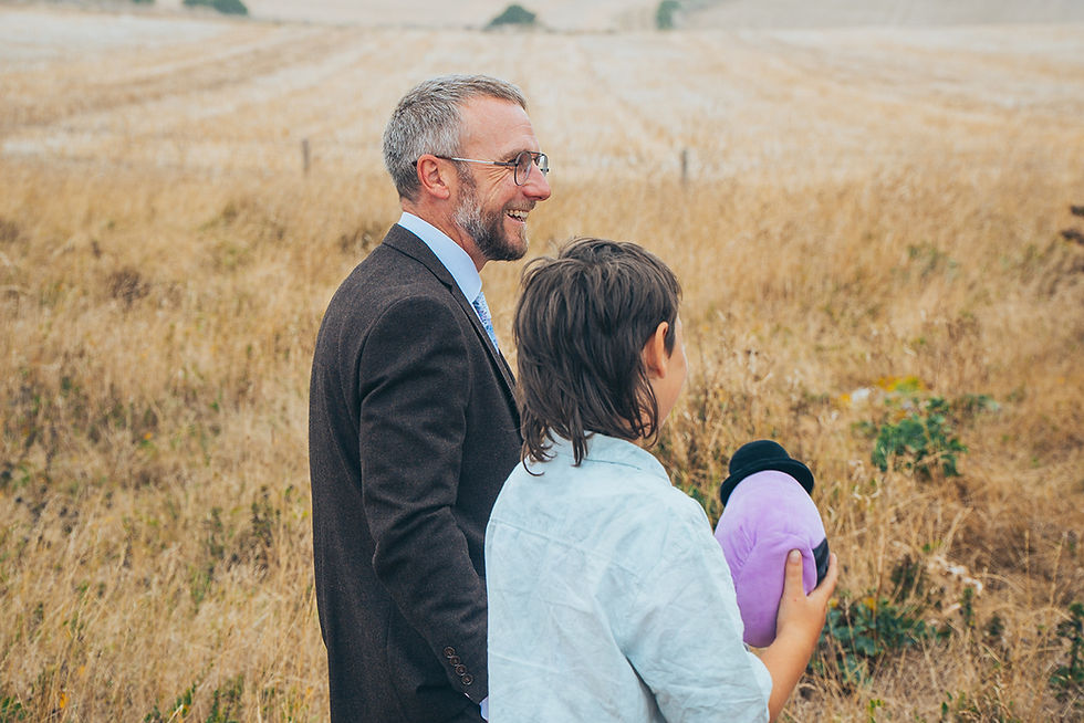 Groom smiling with child holding toy outdoors in Dorset countryside, candid wedding guest moment
