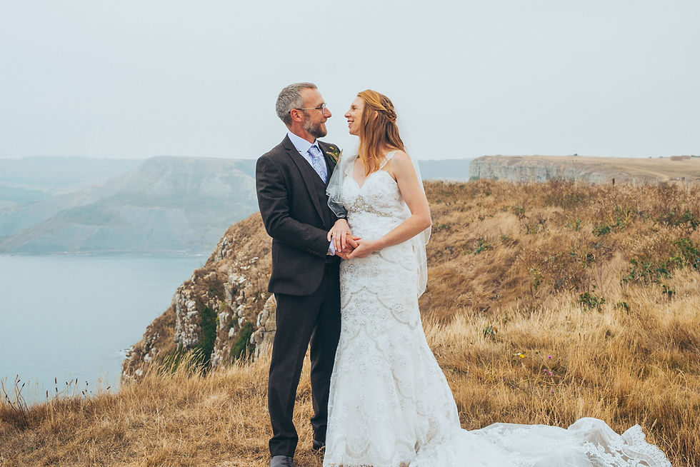 Wedding couple standing on a Dorset clifftop, holding hands and smiling at each other in a natural, candid moment.