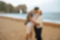 A couple kisses on a pebbled beach with waves in the background. Behind them is a large rocky arch extending into the sea under a cloudy sky.