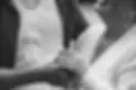 A close-up black and white photo of a smiling couple, focusing on their hands as the woman shows off an engagement ring while gently holding the mans hand. Both wear necklaces and casual clothing.