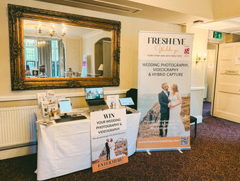 Fresh Eye Weddings display setup at a Spiral Events wedding fayre, featuring elegant signage, wedding photo samples, and a cinematic video reel promoting The Chronicle hybrid photography and videography collection.