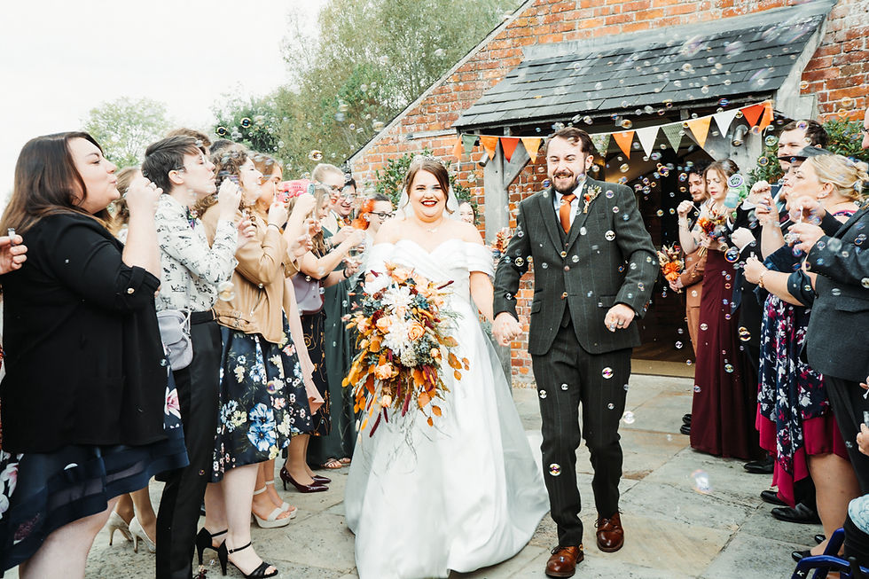 Newly wed couple walking through a joyful bubble confetti exit, surrounded by smiling guests outside a rustic red brick wedding venue in Dorset.
