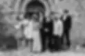 A group of seven people stand in front of a stone building, dressed in formal and semi-formal attire, smiling and talking to each other in a relaxed, celebratory atmosphere.