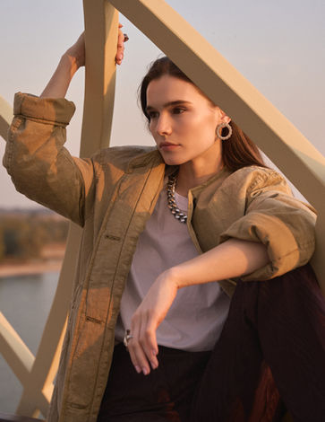 Woman in a khaki jacket, chain necklace and hoop earrings leaning against a wooden lattice at sunset, lake behind