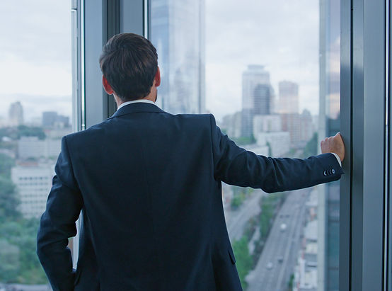 A high achieving, c-suite professional man in a dark suit looking through the glass bracing himself on the wall