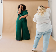 Image of a woman with blonde hair in a white blouse and blue jeans taking a picture of a woman posed in front of a backdrop
