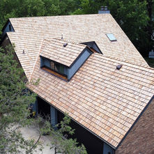 A house with a light brown shingle roof is surrounded by lush green trees. A driveway is visible, and a sign is partially seen in the background.