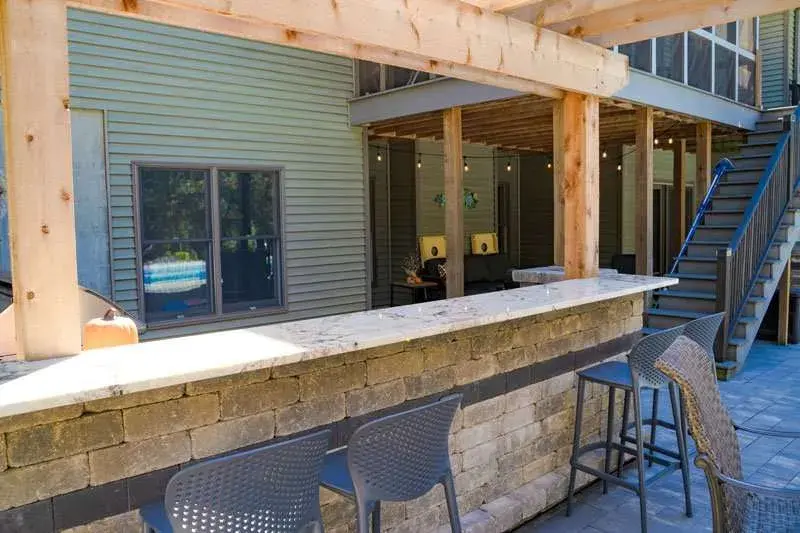 Outdoor patio with stone bar, four gray chairs, and string lights. Open space leads to stairs. Wood pergola casts shadows. Relaxed vibe.