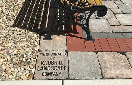 Patio with a park bench and rocks visible with a patio brick engraved with "Patio donated by Knob Hill Landscape Company" in the foreground.
