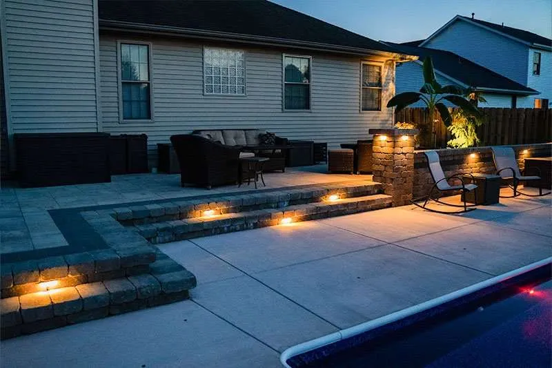 A modern residential patio space in a backyard with a large inground pool and flowering landscaping lining a fence. Space designed by Knob Hill Landscape Company of Springfield, Illinois. 