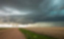 Storm clouds rolling in over a dirt country road and a large expanse of empty fields.