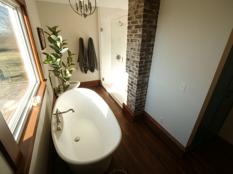 Modern bathroom with exposed brick, a white tub, and large window. Plant beside the tub. Space designed by Capitol Kitchen and Bath in Springfield, Illinois.