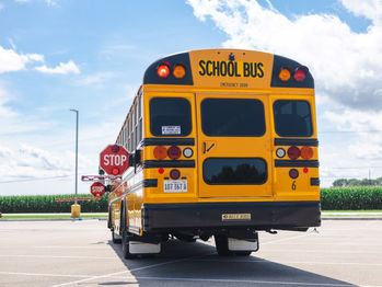 Yellow school bus with "SCHOOL BUS" text, extended stop sign, parked in a lot. Background: blue sky, clouds, and green cornfield.
