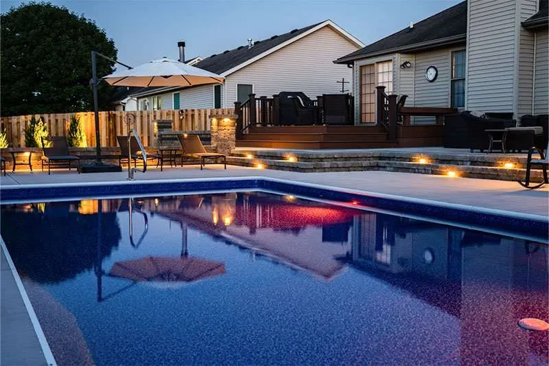 A modern residential patio space in a backyard with a large inground pool and flowering landscaping lining a fence. Space designed by Knob Hill Landscape Company of Springfield, Illinois. 