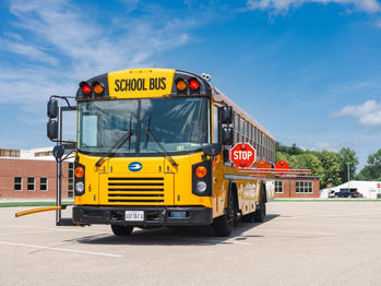 Yellow school bus with a "STOP" sign extended, parked in a lot near a red brick building under a blue sky with clouds.