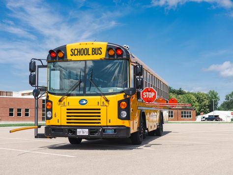 Yellow school bus with a "STOP" sign extended, parked in a lot near a red brick building under a blue sky with clouds.