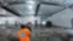 Workers in a large industrial space polish a concrete floor. One wears a bright orange shirt with "Rescue Concrete" text. Ceiling ducts visible.