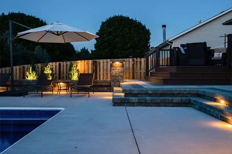 A modern residential patio space in a backyard with a large inground pool and flowering landscaping lining a fence. Space designed by Knob Hill Landscape Company of Springfield, Illinois. 