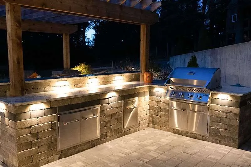 Night view of a backyard patio with built in countertops and grill with a pergola visible in the background. Space designed by Knob Hill Landscape Company of Springfield, Illinois. 