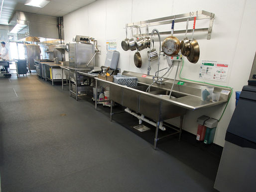 Commercial kitchen with stainless steel appliances, hanging pots, and sink. A chef works at a distant counter. Bright, clean, and organized. ECO Grip flooring installed by Rescue Concrete in Springfield, IL.
