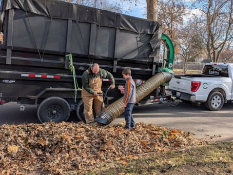 Two people collect leaves with a large vacuum hose attached to a truck on a suburban street. Bare trees and houses in the background.