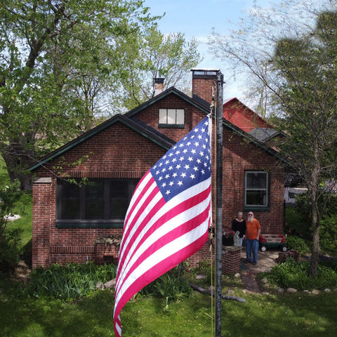 Couple standing on their porch, attached to a brick house, with an American flag in the foreground middle.