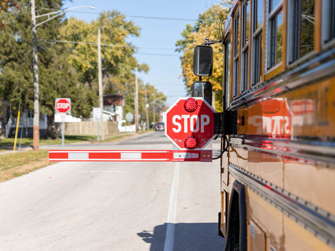 School bus with extended stop sign arm on suburban street; trees and houses line the background; bright, clear day.
