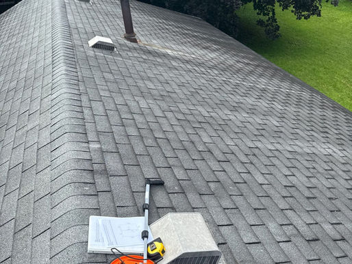 Orange measuring wheel, tape measure, and documents on a grey shingled roof. Background shows trees and green lawn.