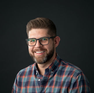 A man with glasses wearing a multi colored checkered button up shirt on a dark grey background.
