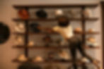 Woman on a stool adjusts hats on wooden shelves in a warm-lit boutique display
