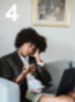 Black woman with afro sits on a couch using a laptop and pen, looking thoughtful.