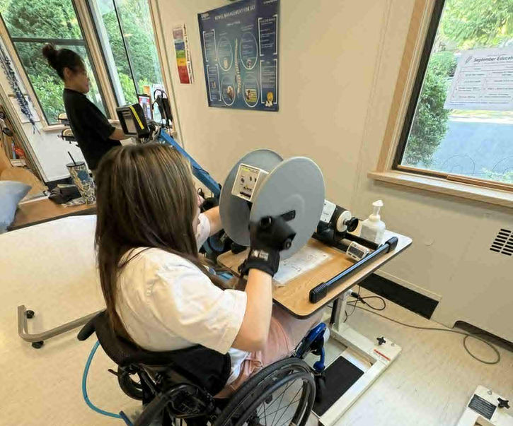 Brook Aleksic in a wheelchair using adaptive exercise equipment inside a rehab center.