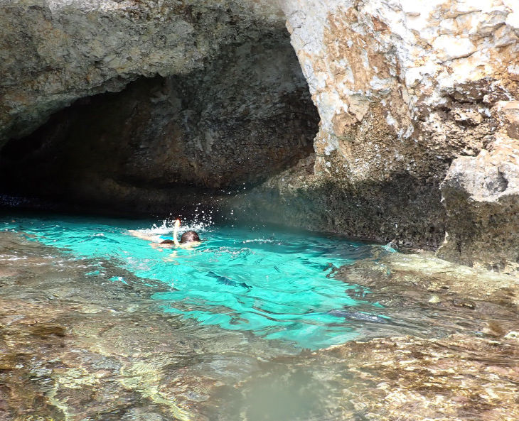 A shot from above sea level. A natural pool with white sand surface surrounded by rocks, at Ayia Napa Sea Caves. A female snorkeler can be seen in the distance, snorkeling towards us.