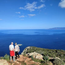 2 walkers in short sleeves standing on top of Moutti tis Sotiras peak looking towards the blue sea under a blue sky in Akamas