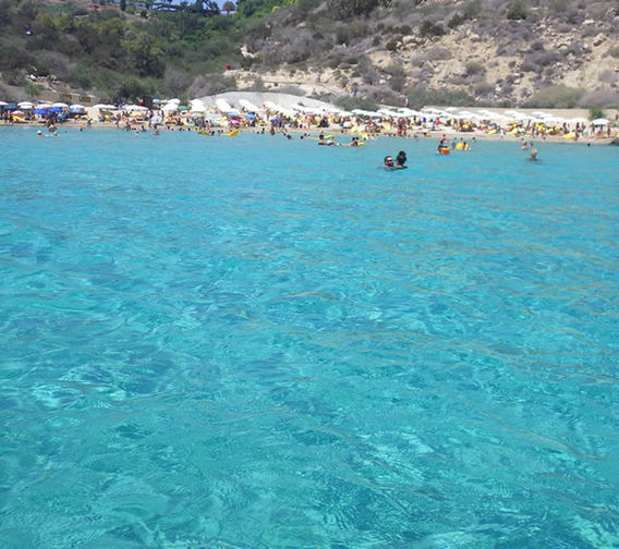 The beautiful calm light blue water of Konnos beach in the foreground. The beach and surrounding hills at the north end of Cape Greco park, far in the background.