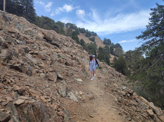 2 lady walkers approaching on Artemis trail, near the top of Olympus peak in Troodos. Barren rock in the foreground, juniper and black pines in the background, some clouds in the sky.
