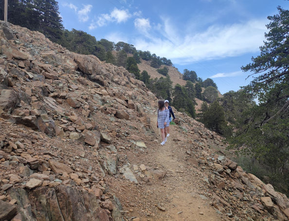 2 lady walkers approaching on Artemis trail, near the top of Olympus peak in Troodos. Barren rock in the foreground, juniper and black pines in the background, some clouds in the sky.