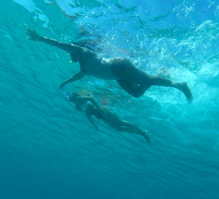 A shot from below from a distance of two female snorkelers at Konnos beach. The water is transparent and the sea surface colour from below is light blue.