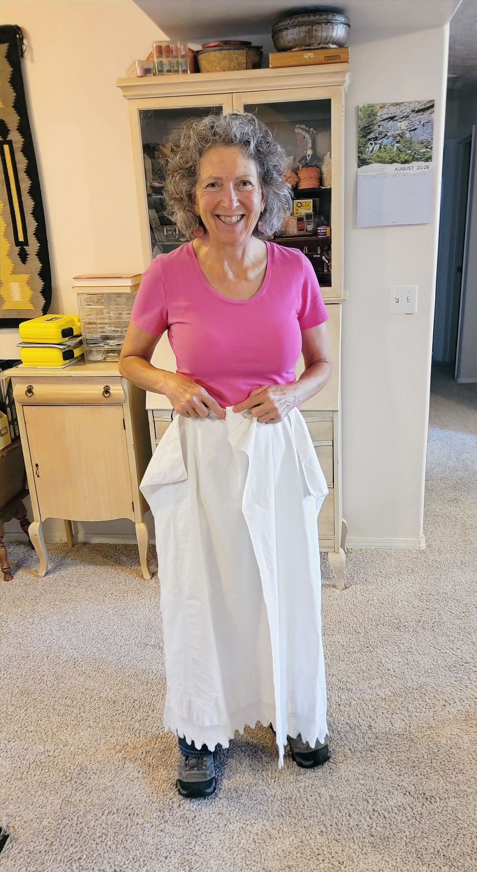 Smiling woman in a pink shirt holds up a white long skirt in a cozy room with a cabinet and August 2026 calendar.