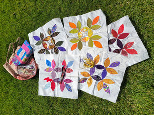 Colorful leaf-pattern quilt blocks and sewing supplies laid on grass; a tote labeled Cynthia sits beside them.