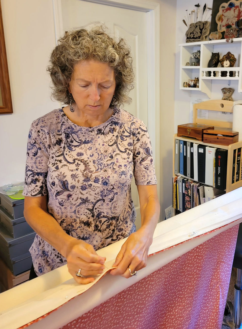 Woman with curly hair carefully sewing fabric at a table in a cozy craft room, with shelves of supplies behind her.