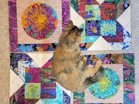 Fluffy cat lies on a bright patchwork floral quilt on beige carpet, surrounded by vivid pink, blue, purple, and green fabric blocks.