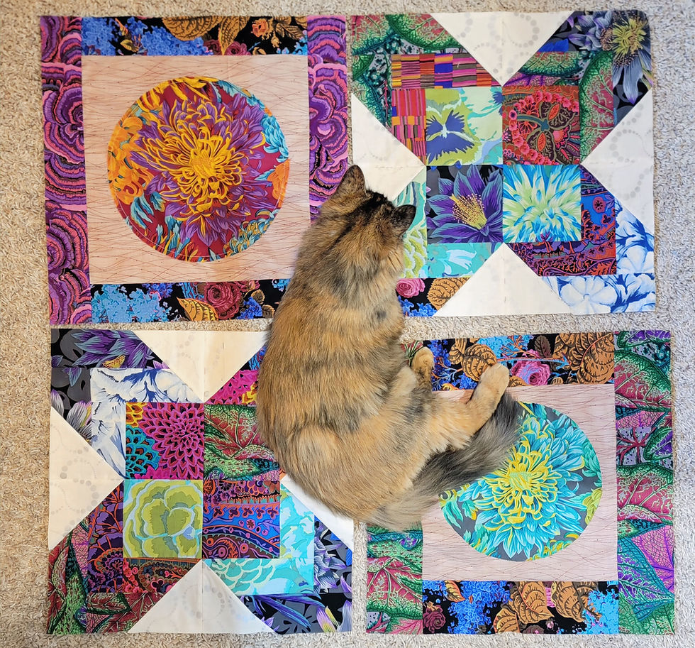Fluffy cat lies on a bright patchwork floral quilt on beige carpet, surrounded by vivid pink, blue, purple, and green fabric blocks.