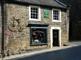 Stone building with a sign reading "The Oldest Sweet Shop in England." A vintage bike decorates the wall. Set in sunlight, year "1661" above door.